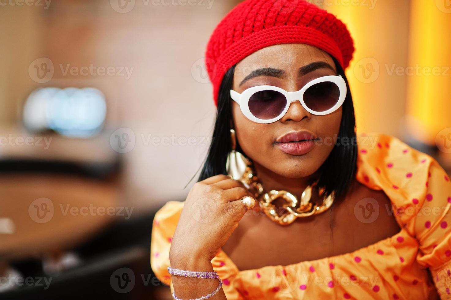 elegante mulher afro-americana na boina vermelha vermelha, grande corrente de pescoço de ouro, óculos de sol, pose de blusa de bolinhas interior. foto