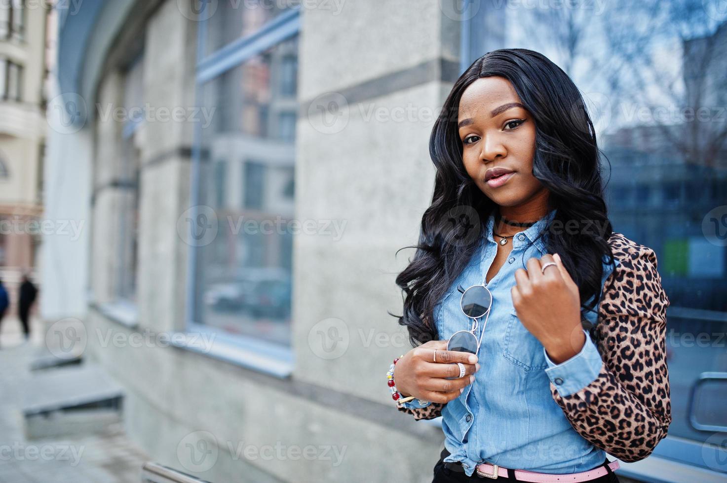 garota afro-americana hipster vestindo camisa jeans com mangas de leopardo posando na rua contra o prédio de escritórios moderno com janelas azuis. foto