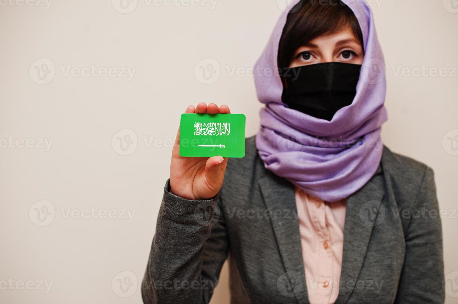 retrato de jovem muçulmana vestindo roupa formal, proteger máscara facial e lenço de cabeça hijab, segurar o cartão de bandeira da arábia saudita contra fundo isolado. conceito de país coronavírus. foto