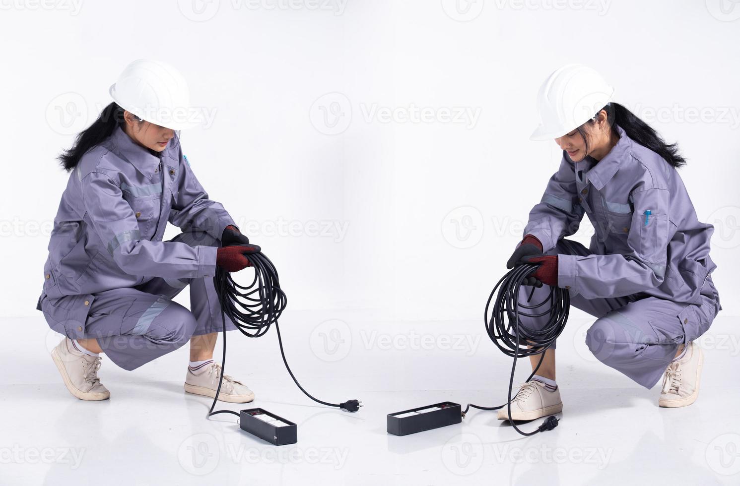 comprimento total 20s jovem mix race engenheiro eletricista mulher, sentado trabalhando duro, usar uniforme cinza. empreiteiro feminino segure o cabo da tomada elétrica sorria feliz sobre fundo branco isolado foto