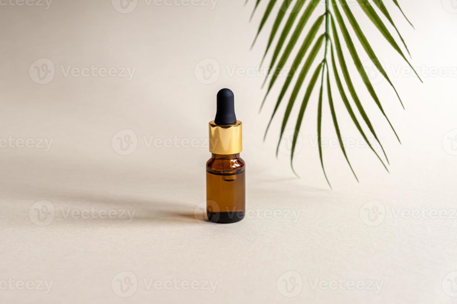 um frasco cosmético de vidro com um conta-gotas em um fundo cinza com folhas de palmeira. conceito de cosméticos naturais, óleo essencial natural foto