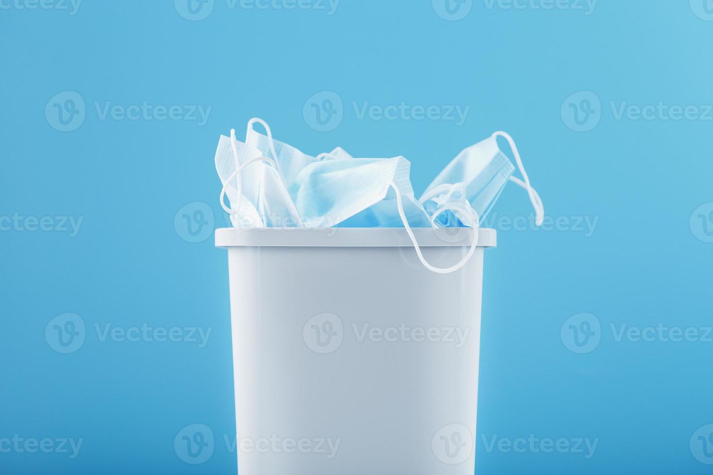 uma lata de lixo branca cheia de máscaras de proteção contra vírus em um fundo azul. foto