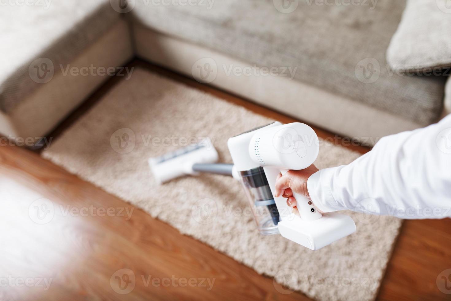 poderoso aspirador sem fio com tecnologia de coleta de poeira ciclônica branca na mão, limpa o tapete da casa perto do sofá. foto