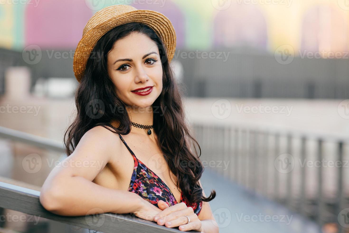 linda mulher com cabelos longos escuros, aparência atraente usando chapéu de verão e vestido tendo excursão na cidade grande posando na ponte. jovem viajante feminina andando na rua tendo um olhar delicioso foto