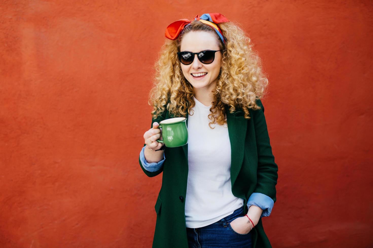 Mulher jovem e atraente europeia com bandana vestindo jaqueta elegante e tons segurando a xícara de cappuccino tendo expressão feliz em pé contra um fundo laranja. pessoas, conceito de estilo de vida foto