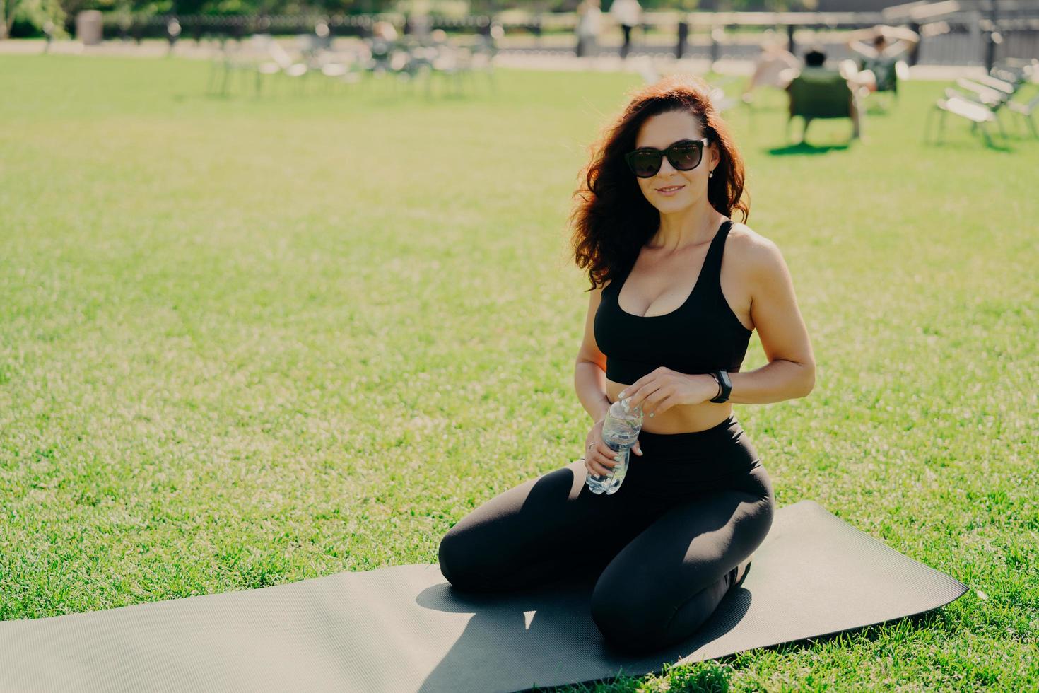 foto de mulher caucasiana desportiva senta-se no tapete bebe água após o exercício sente-se revigorado e satisfeito entra para o esporte ao ar livre durante o dia de verão usa óculos escuros e roupas esportivas. estilo de vida saudável