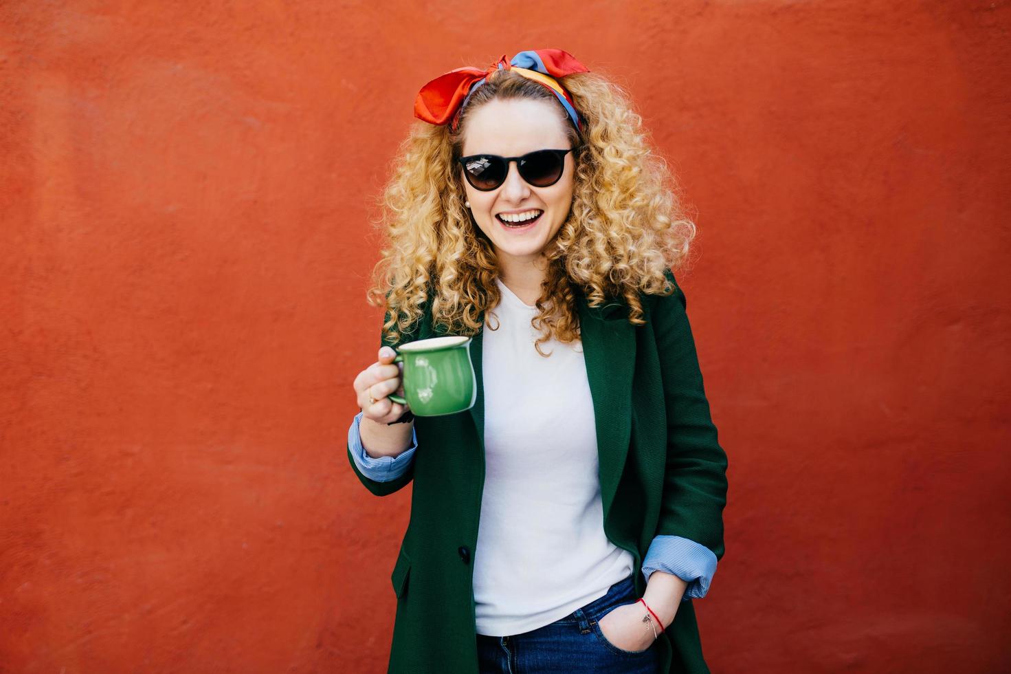 aluna bonita com cabelo loiro encaracolado usando bandana, óculos escuros e elegante jaqueta segurando a xícara de café olhando para a câmera com um sorriso sincero. mulher bonita gostando de beber chá saboroso foto