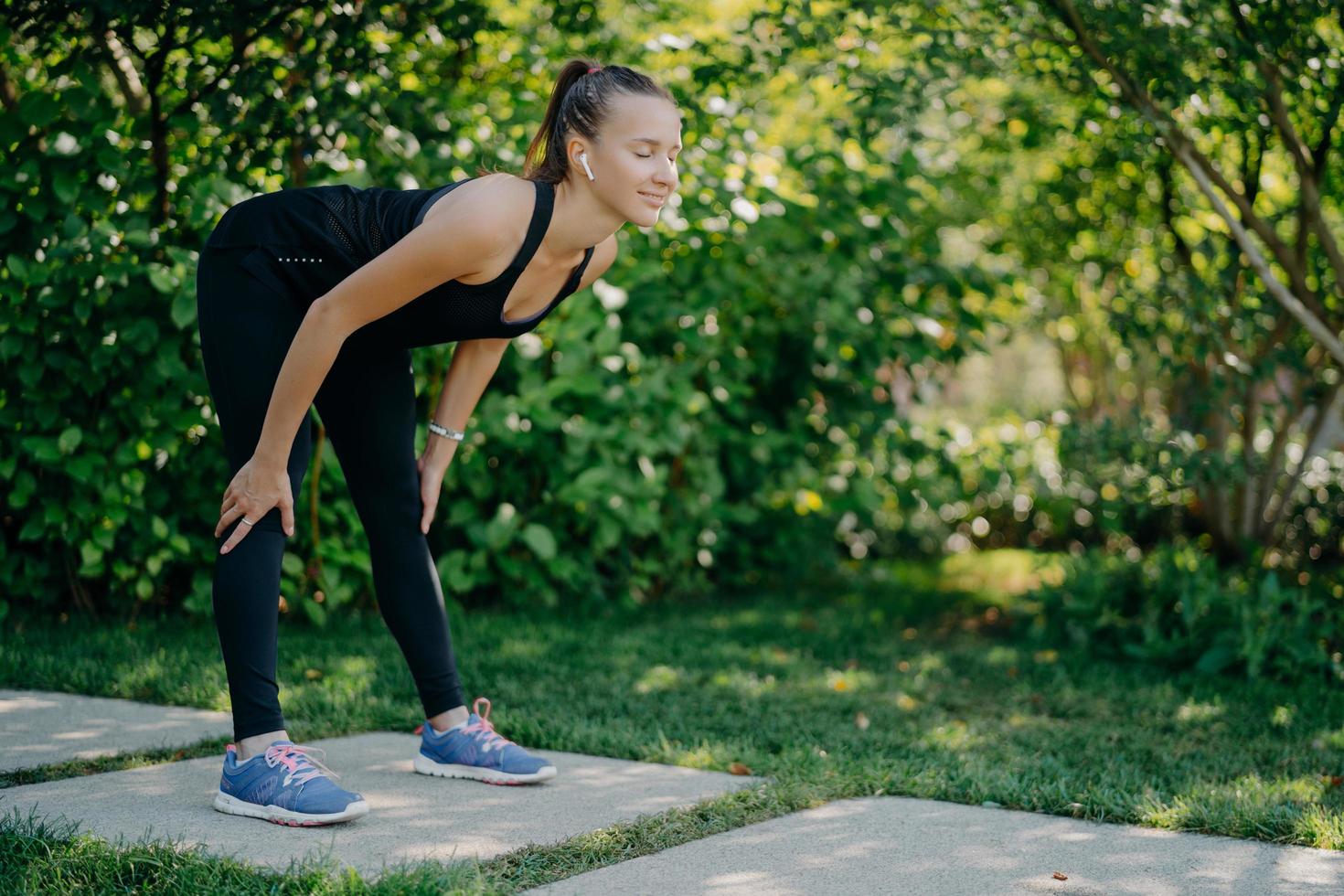 mulher fitness em roupas esportivas pretas mantém as mãos nos joelhos se inclina para a frente exercícios ao ar livre contra o fundo verde da natureza ouve música da lista de reprodução favorita. perda de peso e conceito de treino foto