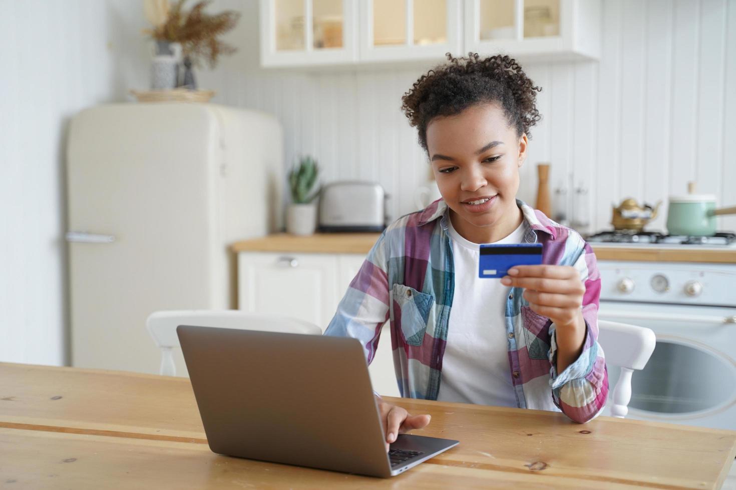 cliente de comércio eletrônico jovem garota biracial usando cartão de crédito bancário para pagar on-line no laptop em casa foto