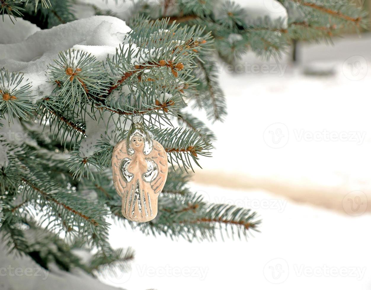 anjo de brinquedo de natal pendura em um galho coberto de neve de uma árvore de natal em um fundo festivo de bokeh de neve branca com espaço de cópia. foto