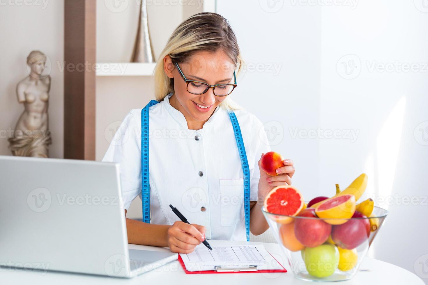 nutricionista sorridente em seu escritório, ela está segurando uma fruta e mostrando vegetais e frutas saudáveis, conceito de saúde e dieta. nutricionista feminina com frutas trabalhando em sua mesa foto