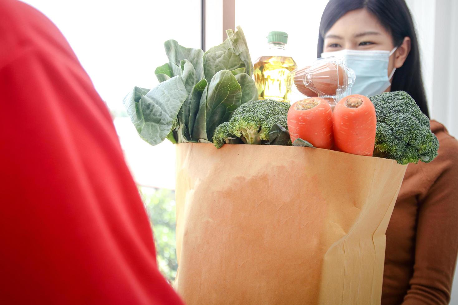 um mensageiro carregando uma sacola de comida, ovos, legumes e ingredientes entregues a uma cliente do sexo feminino usando uma máscara bem na porta da frente. conceito de negócio de serviço de entrega comprando comida online durin foto