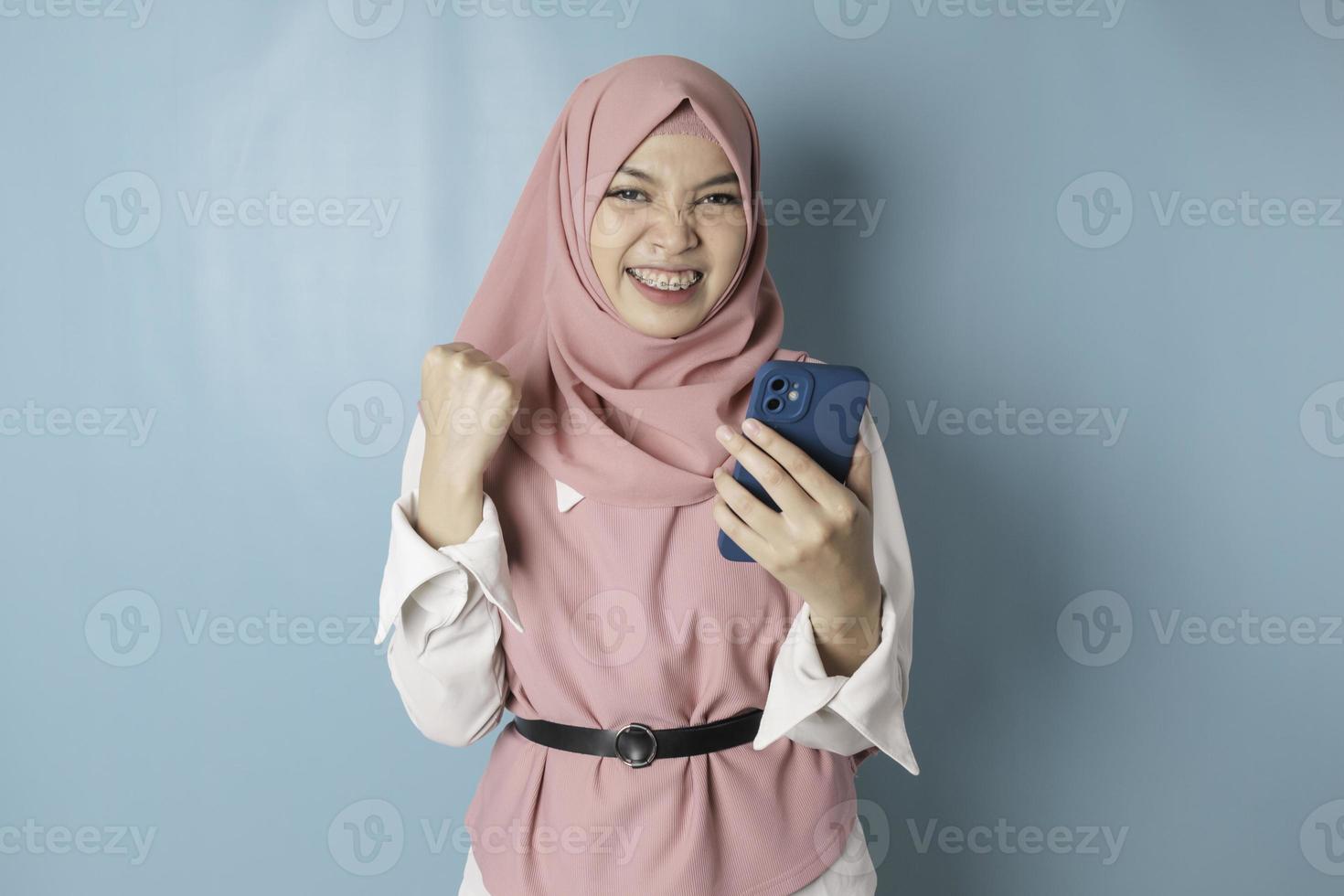 uma jovem muçulmana asiática com uma expressão feliz e bem sucedida usando hijab rosa e segurando smartphone isolado por fundo azul foto