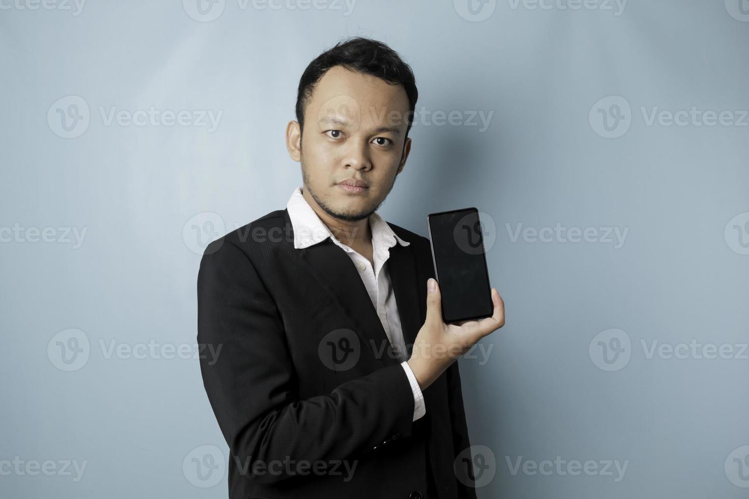 um retrato de um empresário asiático feliz está sorrindo e segurando seu smartphone mostrando espaço de cópia na tela vestindo terno preto isolado por um fundo azul foto