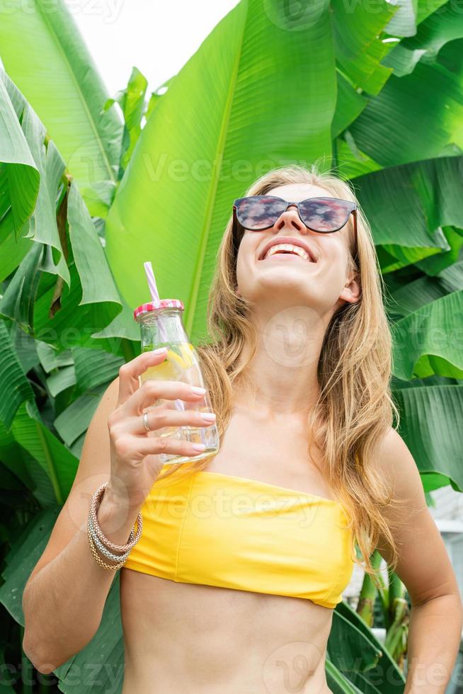 jovem mulher caucasiana em maiô amarelo bebendo bebida em pé em folhas de palmeira tropical foto
