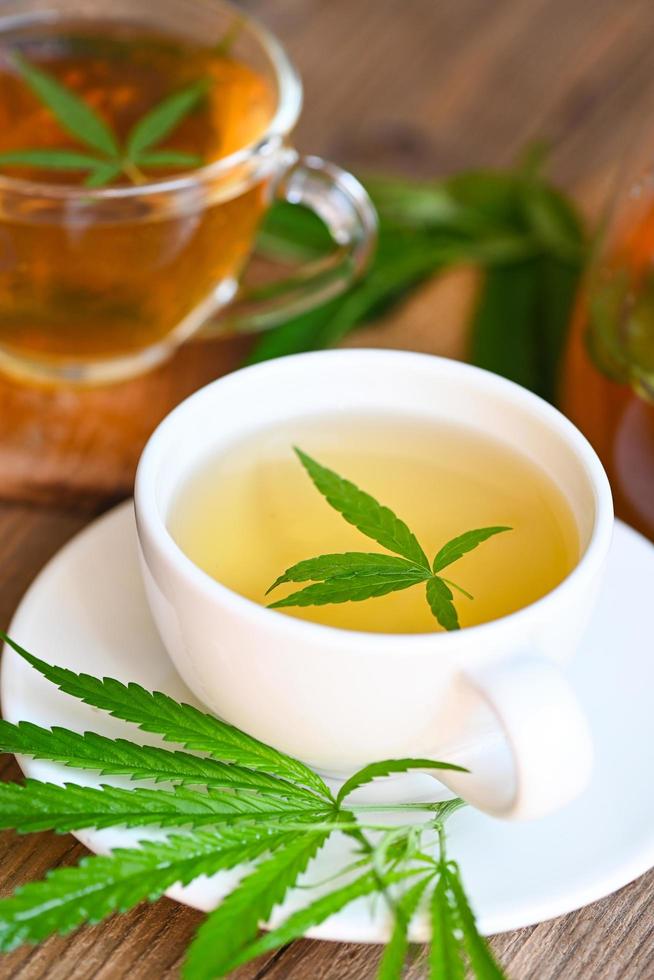 chá de cannabis ervas na xícara de chá com folha de cannabis folhas de maconha erva em fundo de madeira, chá de saúde com planta de folha de cânhamo thc cbd ervas comida e conceito médico foto