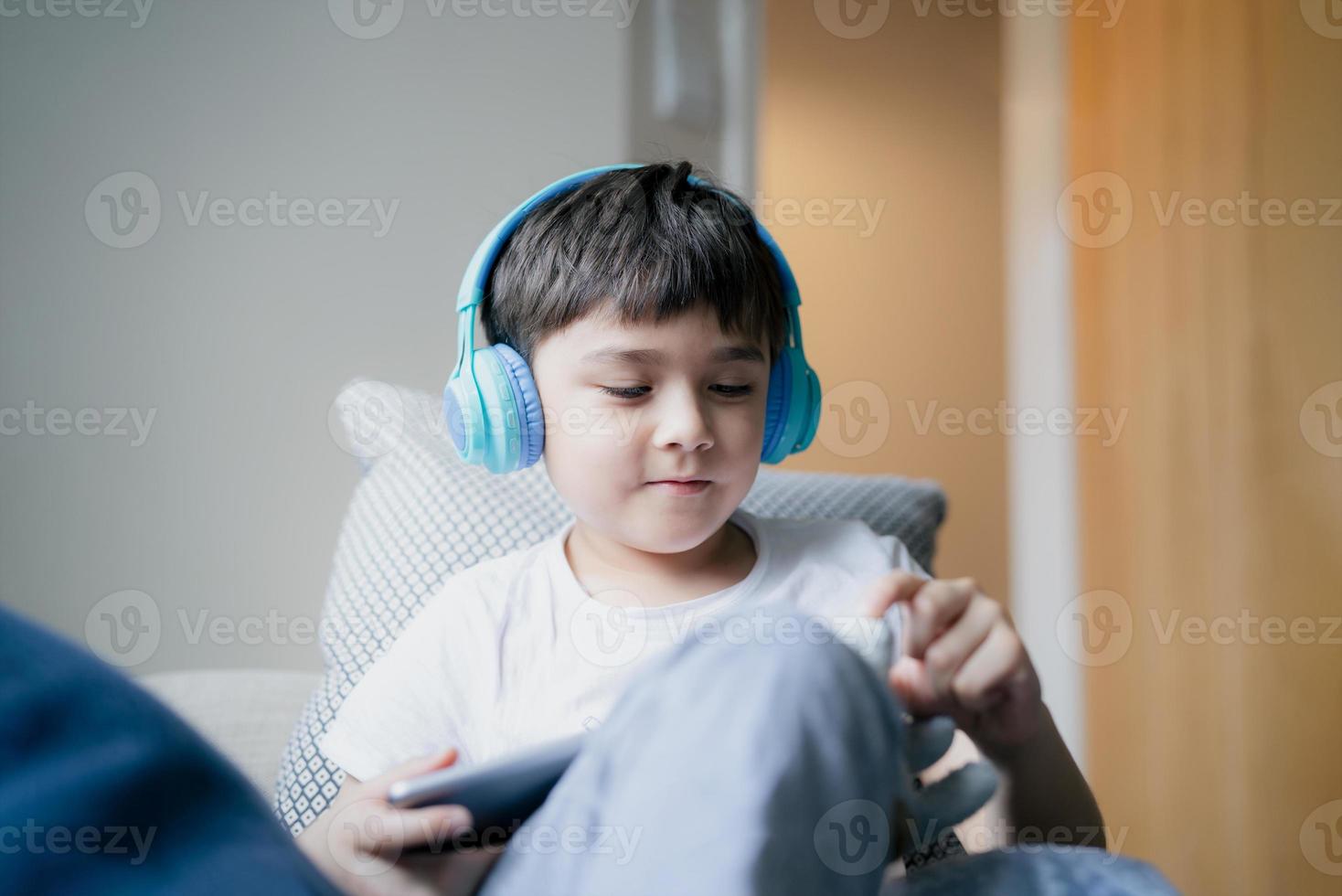 menino feliz usando fone de ouvido para jogar na internet com amigos, criança sentada ao lado da janela lendo ou assistindo desenhos animados no tablet, criança da escola fazendo lição de casa online em casa. foto