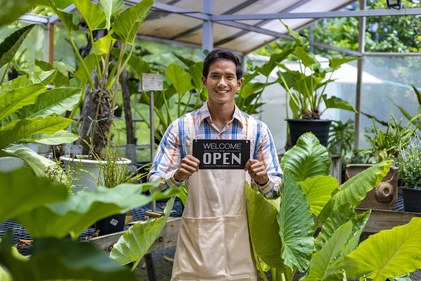 jardineiro asiático segurando placa de abertura para receber o cliente em seu centro de viveiro tropical cheio de espécies exóticas de samambaias foto