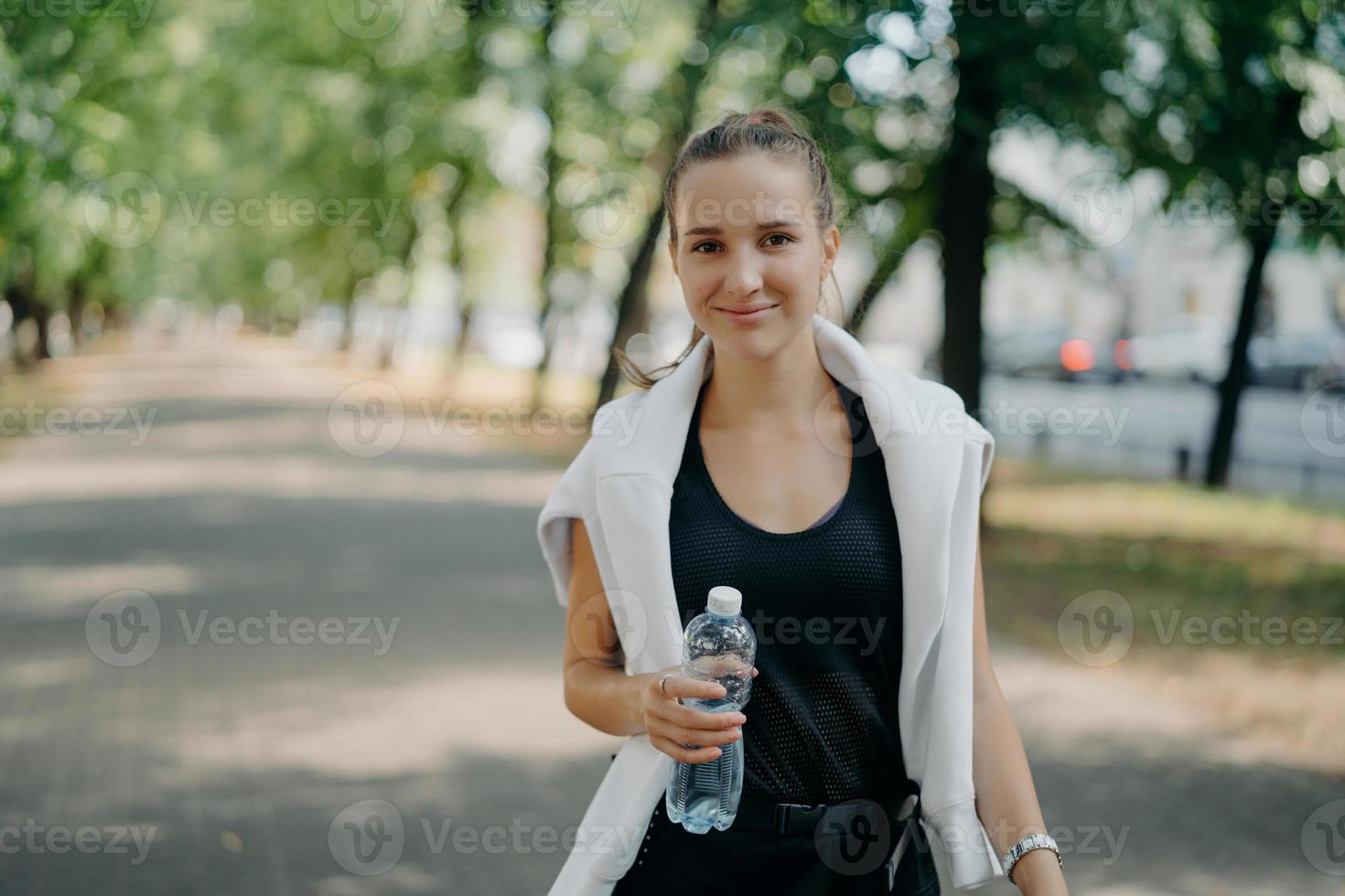 satisfeito magro jovem europeu desportivo envolvido em bebidas desportivas água doce vestida com roupa ativa passeios no parque durante o dia ensolarado tem expressão do rosto satisfeito. hidratação depois do treino cardio foto