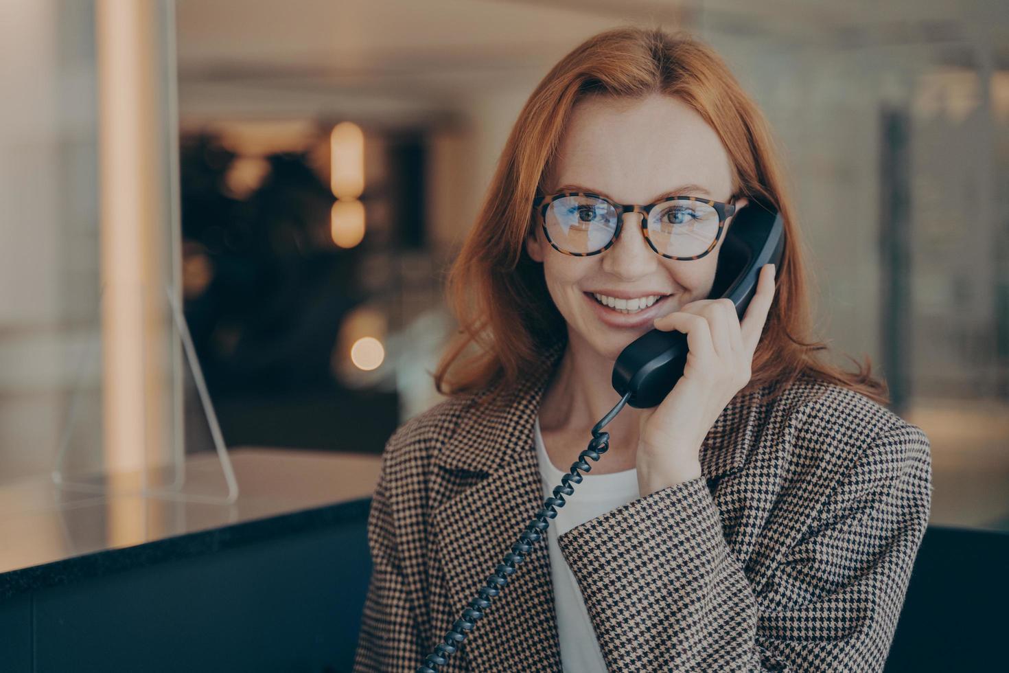 trabalhadora de escritório feminina em óculos falando no telefone fixo com colega de trabalho e sorrindo para a câmera foto