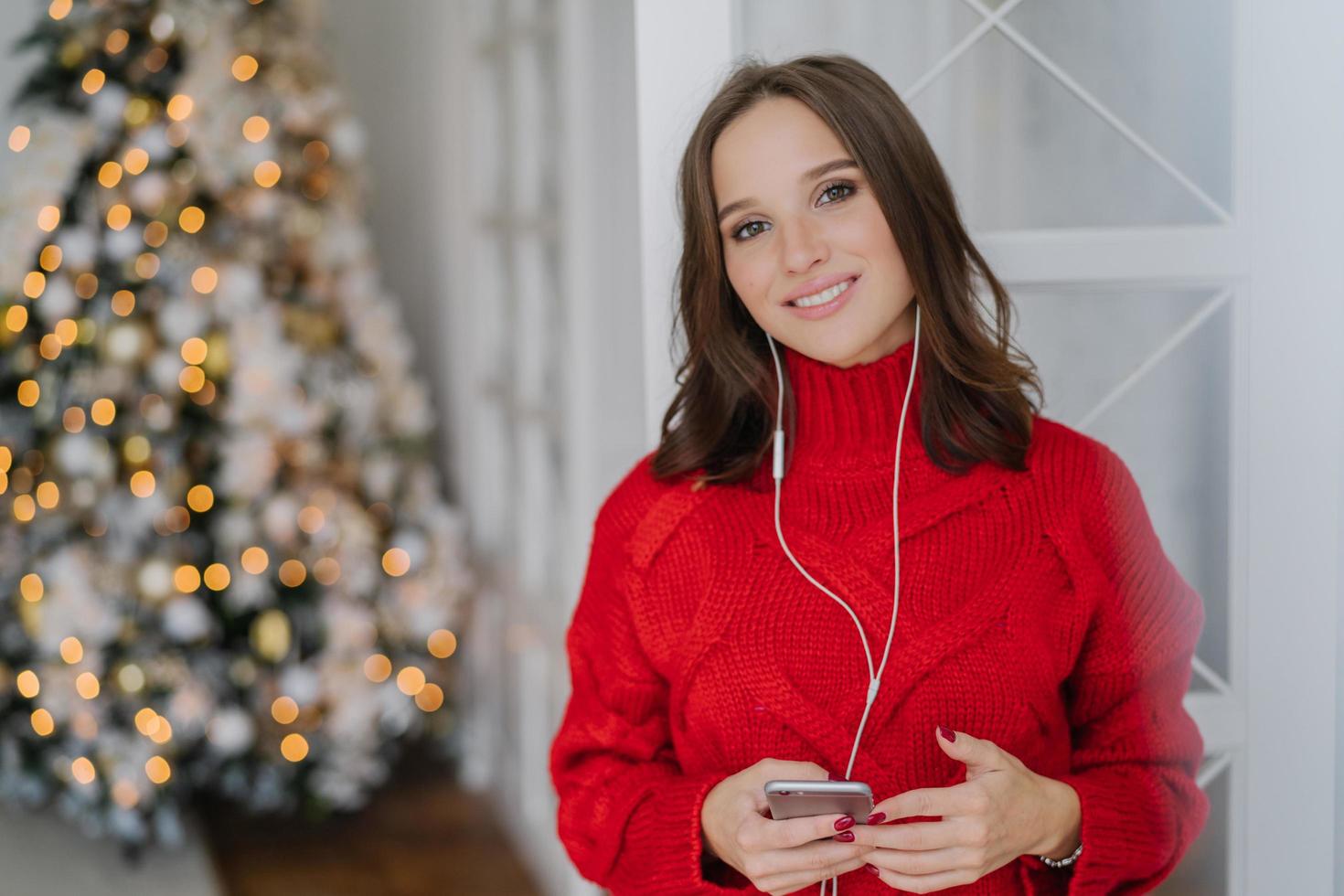 alegre mulher bonita tem sorriso cheio de dentes, gosta de uma boa faixa em fones de ouvido, atualiza a lista de reprodução no telefone inteligente, vestida com um suéter de malha quente, posa contra a árvore de natal decorada em segundo plano foto