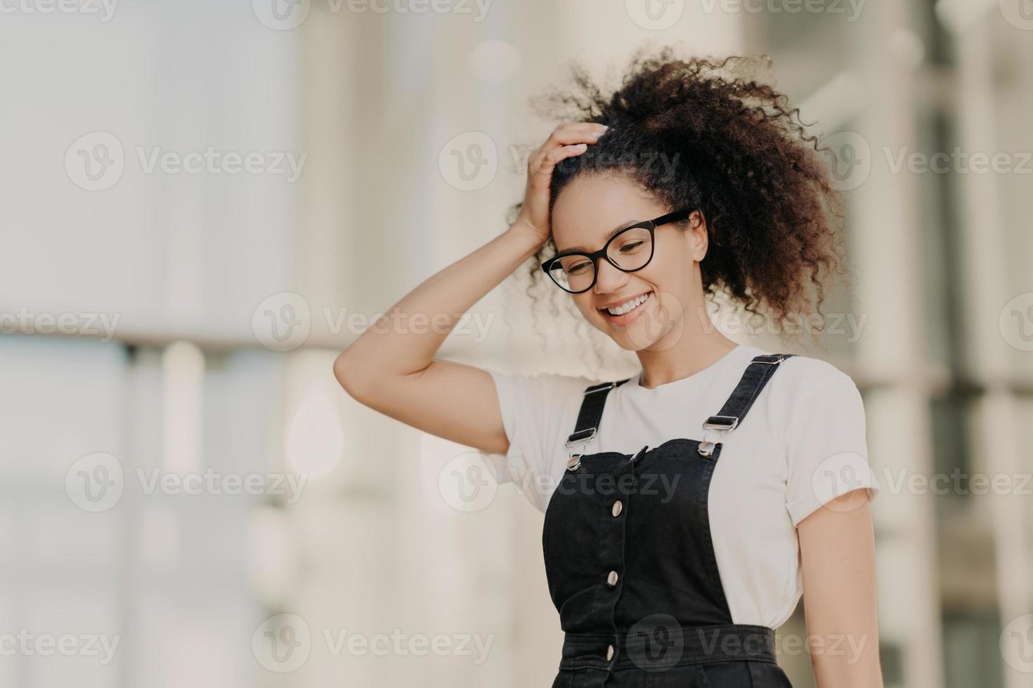 linda modelo feminina de pele escura com cabelos crespos, olha para baixo, vestida com camiseta branca casual, macacão preto, gosta de tempo livre, caminha ao ar livre, satisfeita com boas notícias. boas emoções e pessoas foto