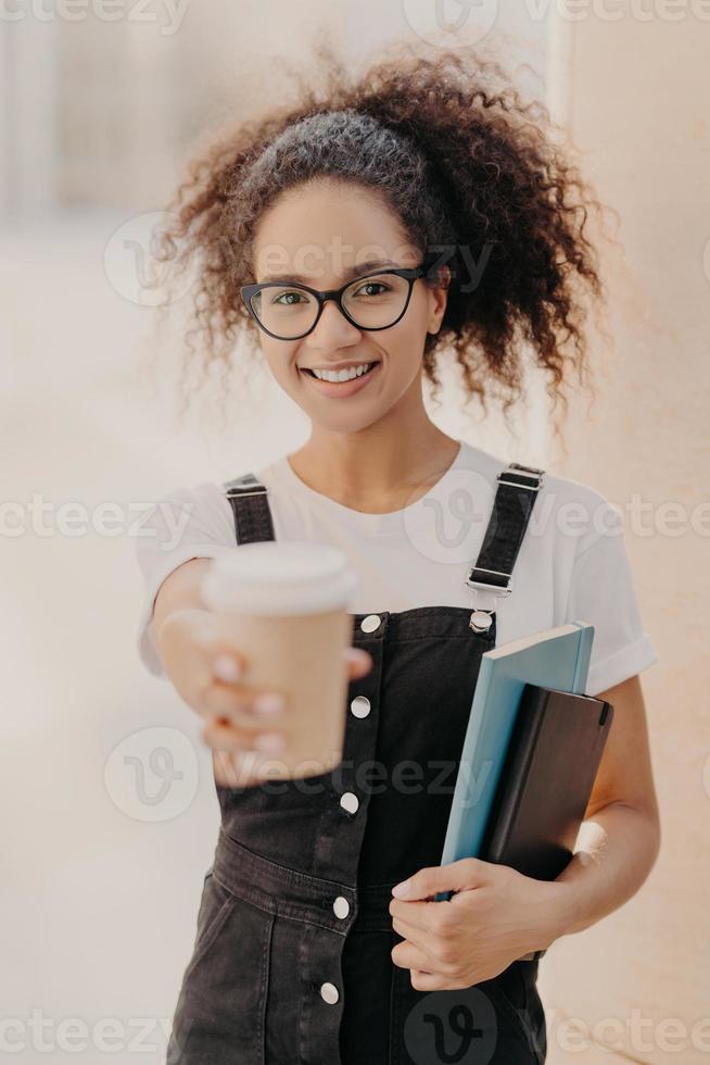 tiro vertical de mulher bonita com cabelos cacheados, segura café para viagem, bebe durante as férias na universidade, usa óculos e macacão jeans, expressa emoções positivas. estudante feliz com bebida foto
