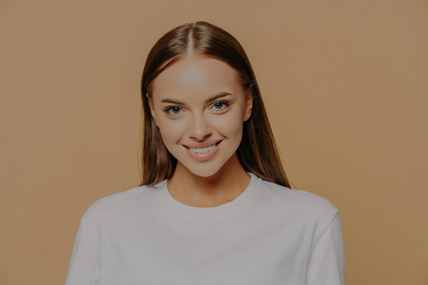 retrato de mulher bonita com sorrisos longos e lisos cabelos escuros suavemente usa jumper branco casual tem poses de maquiagem mínima contra fundo marrom. beleza feminina e conceito de emoções positivas foto