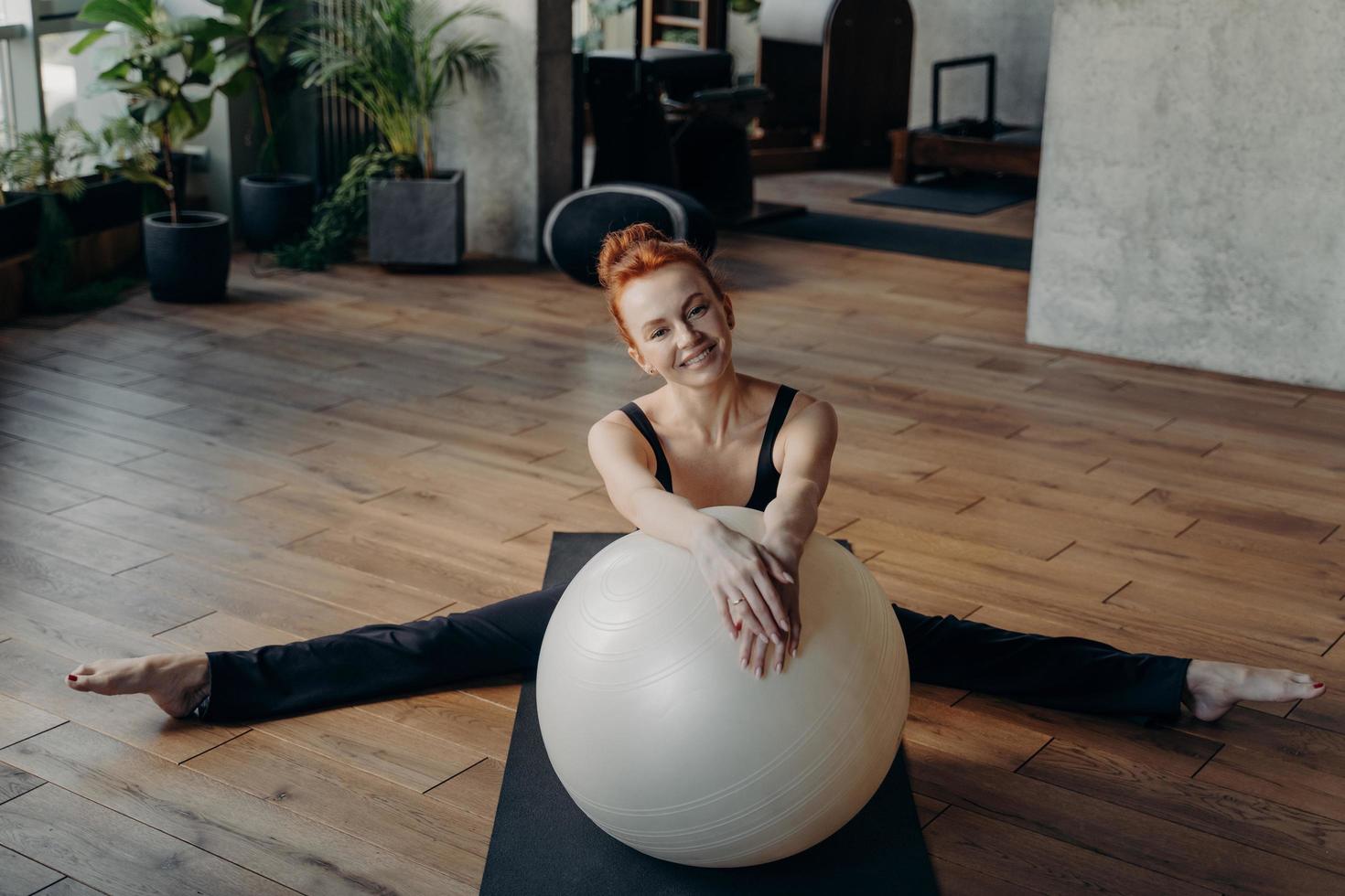 mulher saudável ruiva sorridente em posição dividida, alongando-se com fitball foto