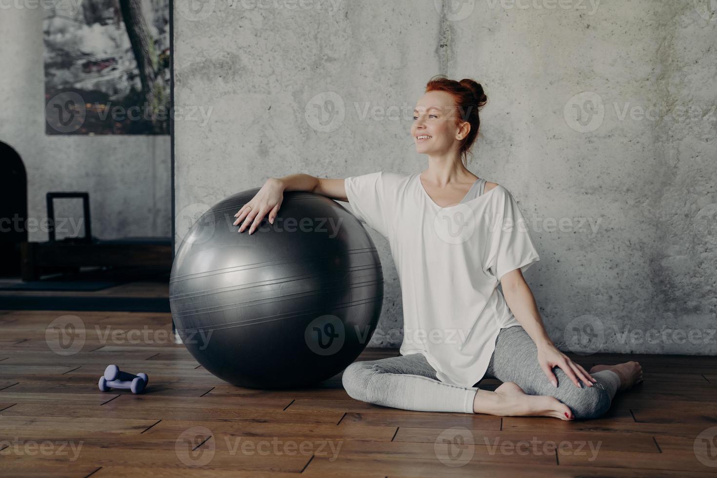 mulher de fitness feliz senta-se elegantemente no chão de parguet, apoiando uma mão na fitball prata foto