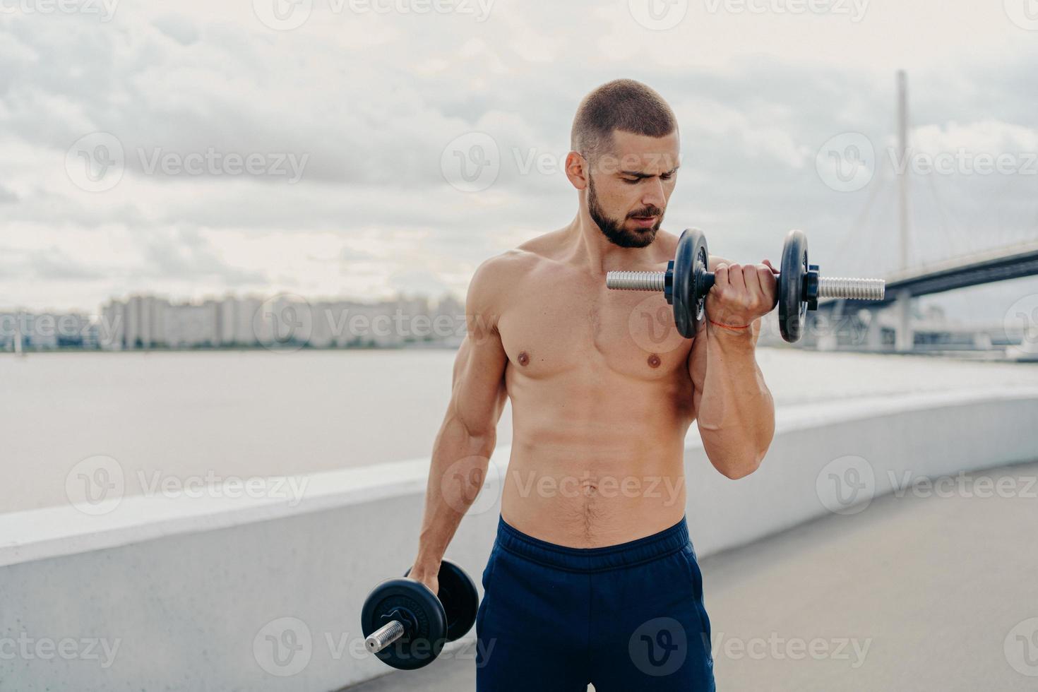 cara musculoso com exercícios de torso nu com halteres, fica sem camisa, treina músculos ao ar livre, fica em piere, tem motivação para se manter em forma e saudável. fitness, esporte, conceito de musculação foto