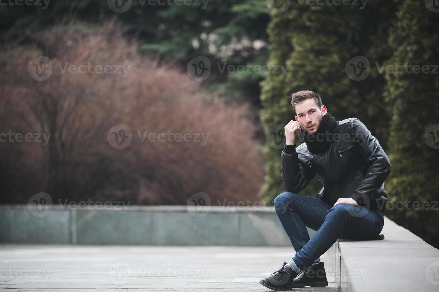 um homem vestido de jeans e assentos de jaqueta preta em uma laje foto