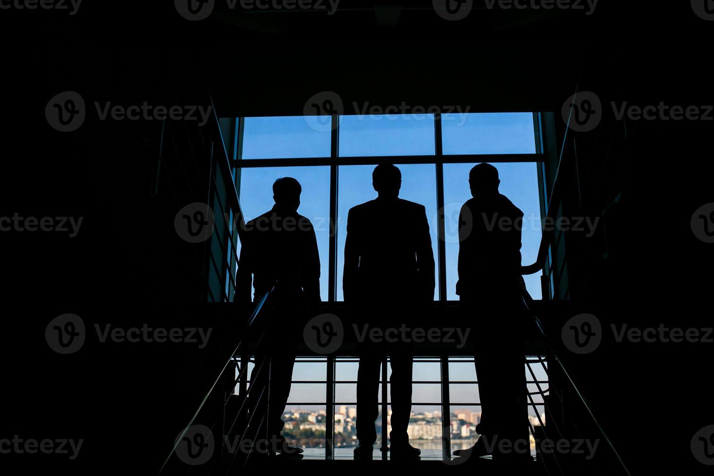 três empresários em pé na janela com vista para a cidade foto