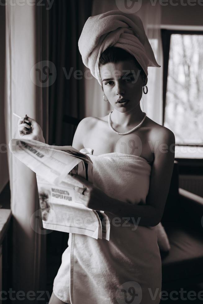 mulher jovem e bonita em uma toalha fuma um cigarro e lê jornal foto