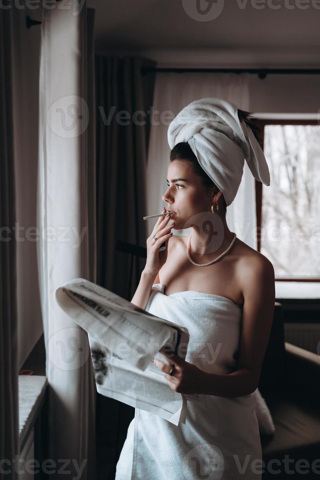 mulher jovem e bonita em uma toalha fuma um cigarro e lê jornal foto