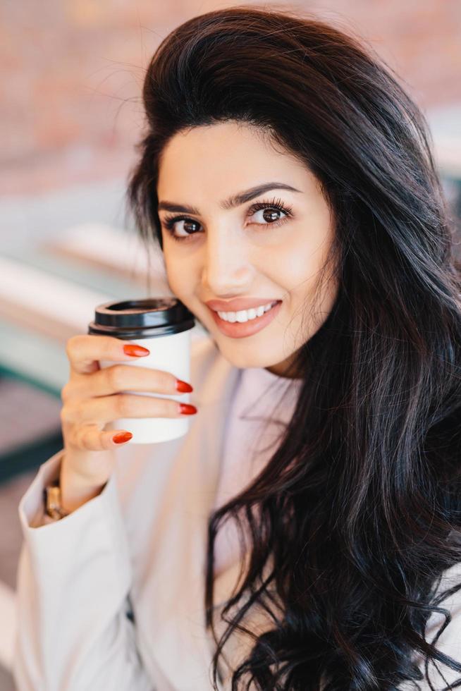 conceito de beleza, juventude, estilo de vida e relaxamento. jovem morena com sobrancelhas finas bem formadas, pele saudável e manicure vermelha segurando café para viagem olhando agradavelmente para a câmera sorrindo foto