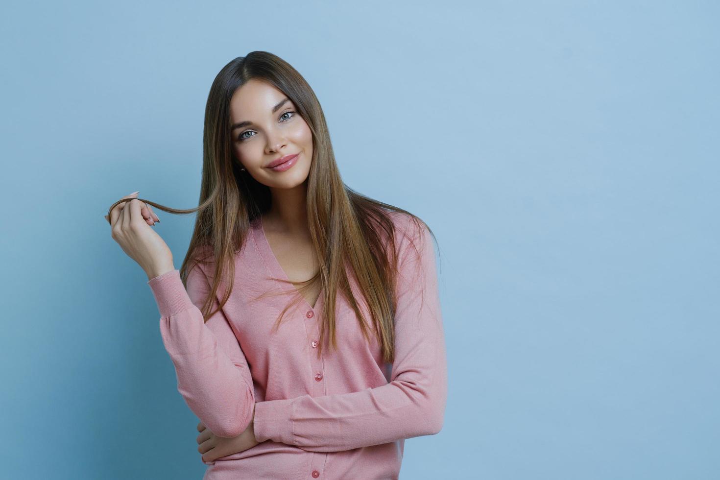 tiro da cintura para cima de uma jovem de aparência agradável tem lindos cabelos longos, inclina a cabeça, vestida com um jumper casual, tem a pele limpa perfeita, anuncia xampu para cuidados com os cabelos, isolado em fundo azul. foto