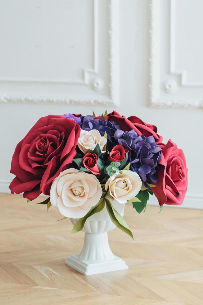 tiro vertical de lindo buquê com rosas artificiais vermelhas e brancas em vaso branco no piso de madeira contra fundo branco. arranjo de flores. fechar-se foto