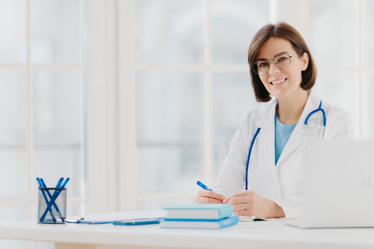 especialista médico usa vestido branco com estetoscópio, trabalha no hospital, faz anotações, posa no próprio gabinete, senta-se à mesa com o laptop no escritório sorri alegremente. cuidados de saúde e conceito médico foto