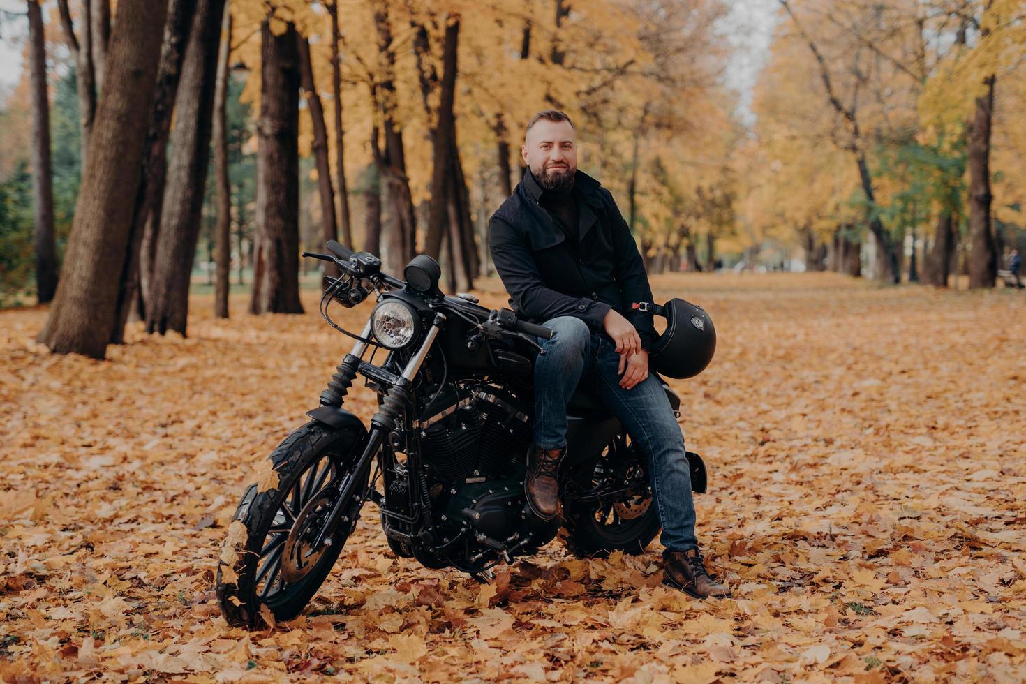 motociclista barbudo posa na própria moto, segura o capacete, anda de moto, posa ao ar livre no parque, chão coberto de folhas caídas. estilo de vida do motociclista. aventura em transporte próprio. cavaleiro masculino ao ar livre foto