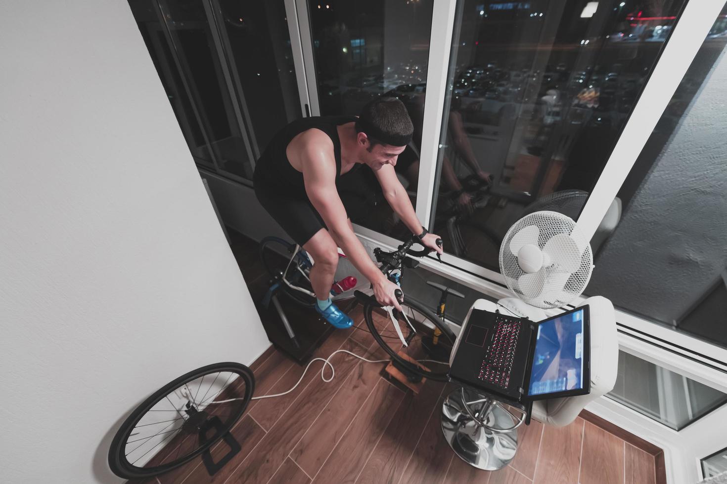 homem andando de bicicleta no treinador de máquina ele está se exercitando em casa à noite jogando jogo de corrida de bicicleta online foto