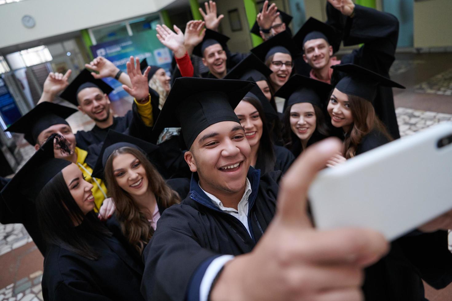 grupo de estudantes internacionais felizes em placas de argamassa e vestidos de solteiro com diplomas tomando selfie por smartphone foto