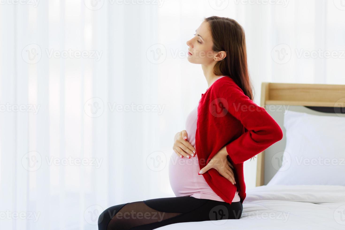 mulher grávida senta-se na cama no quarto, segurando as costas, mostrando dor ou dor nas costas. foto