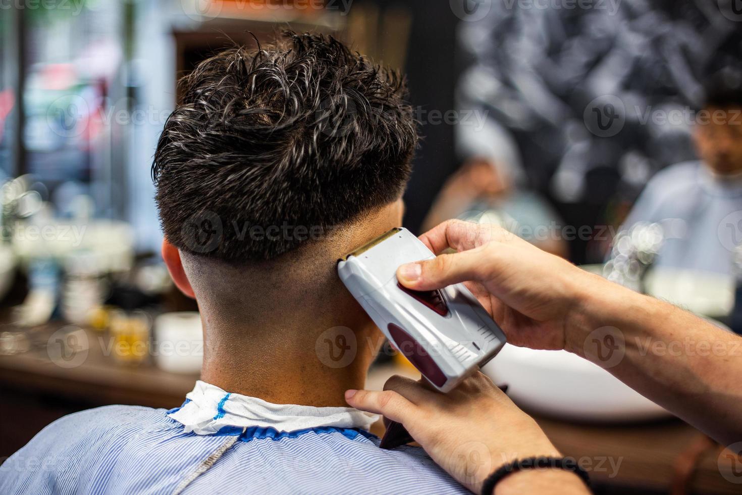 vista traseira do jovem recebendo um corte de cabelo moderno. homem sendo aparado com máquina de tosquiadeira elétrica profissional na barbearia. conceito de tratamento de beleza masculina. cara recebendo novo corte de cabelo no salão de barbeiro foto