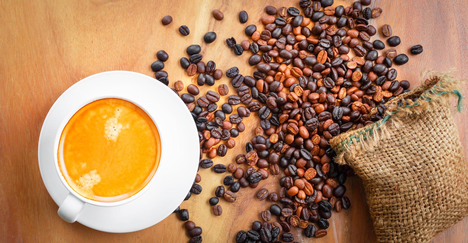 vista superior da xícara de café tem espuma dourada e grãos de café misturados ou misturados em um saco marrom no fundo de madeira foto