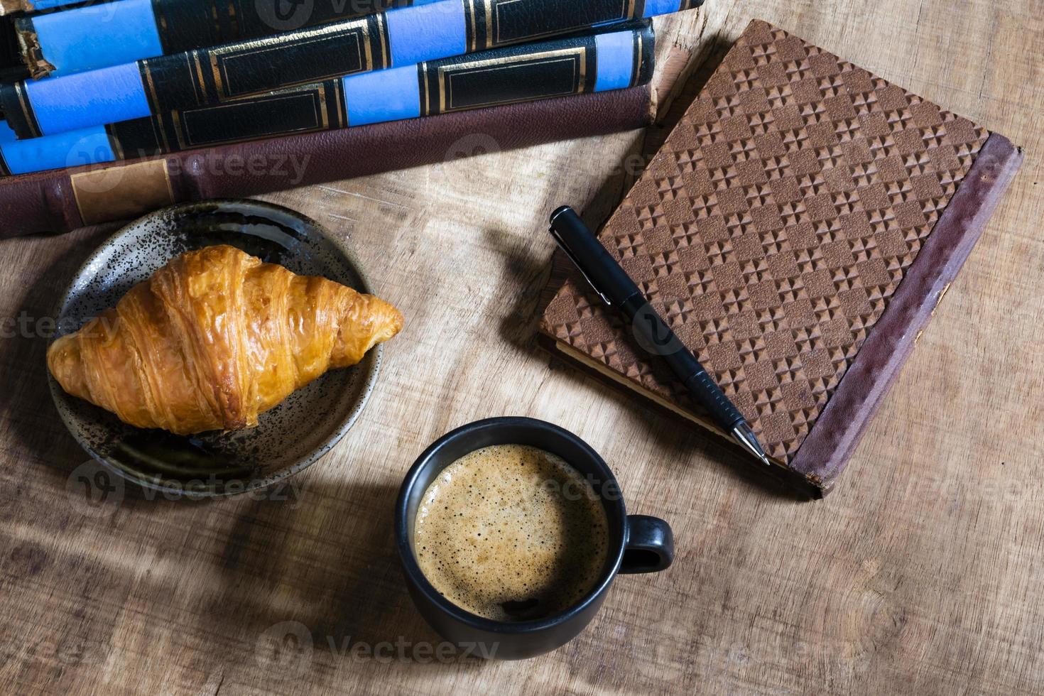 croissant de café da manhã no prato e café tem livro na mesa de madeira. foto