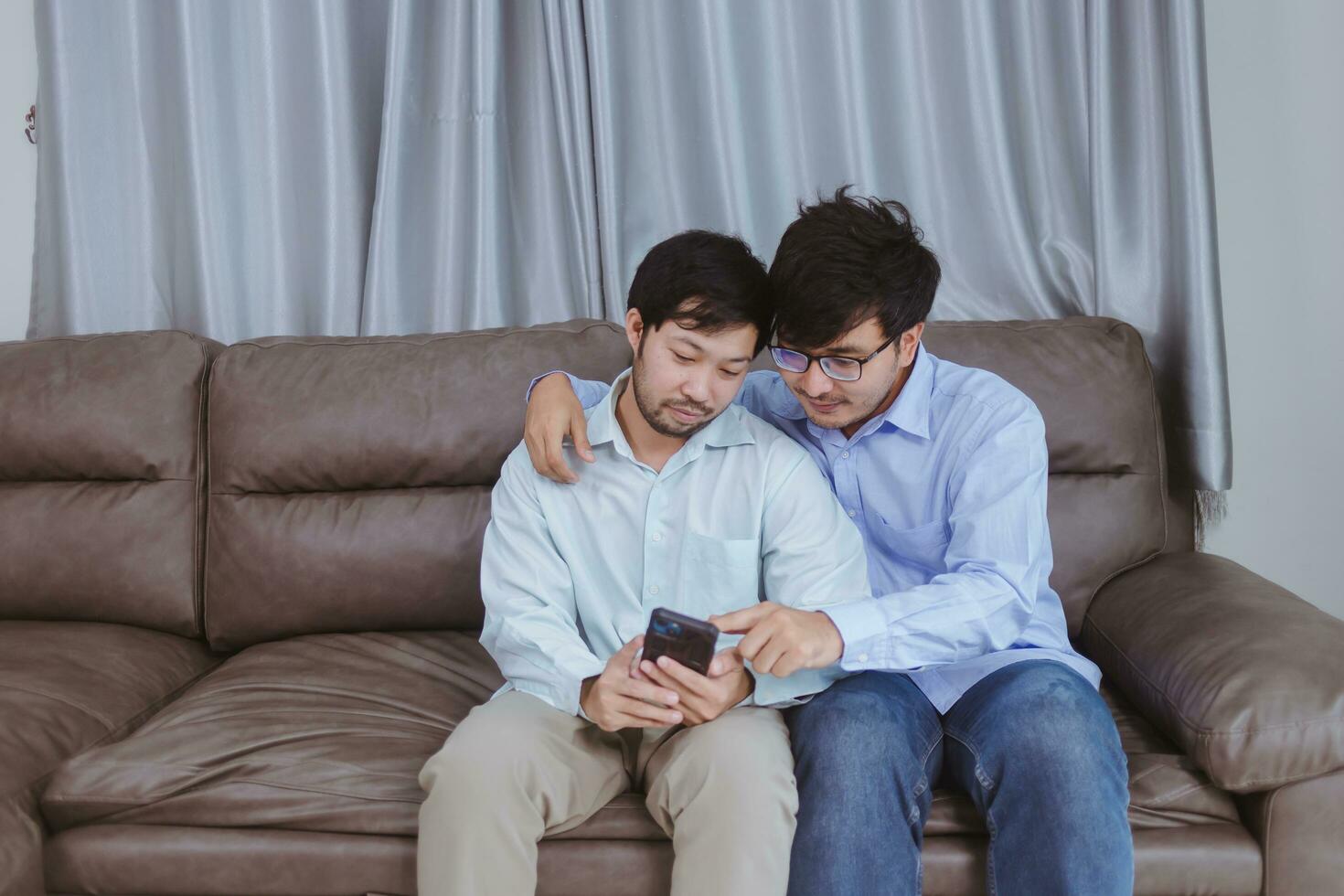 feliz casal gay passando o dia juntos em casa. homem e parceiro olhando para o telefone no sofá. foto