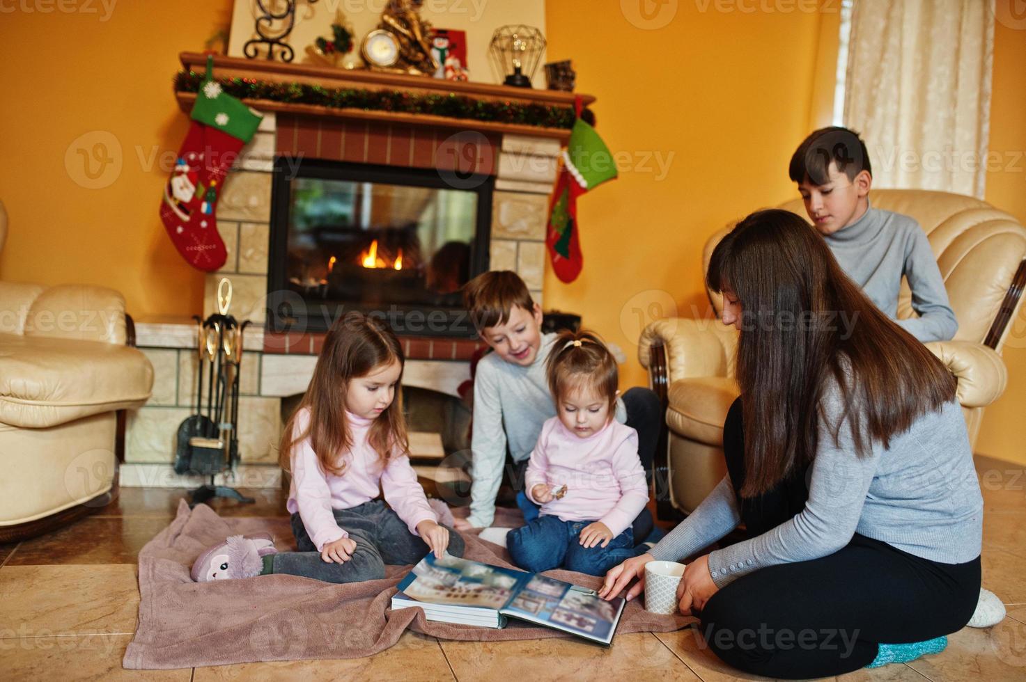 feliz jovem grande família por uma lareira na sala de estar quente em dia de inverno. mãe com quatro filhos em casa leu o livro. foto