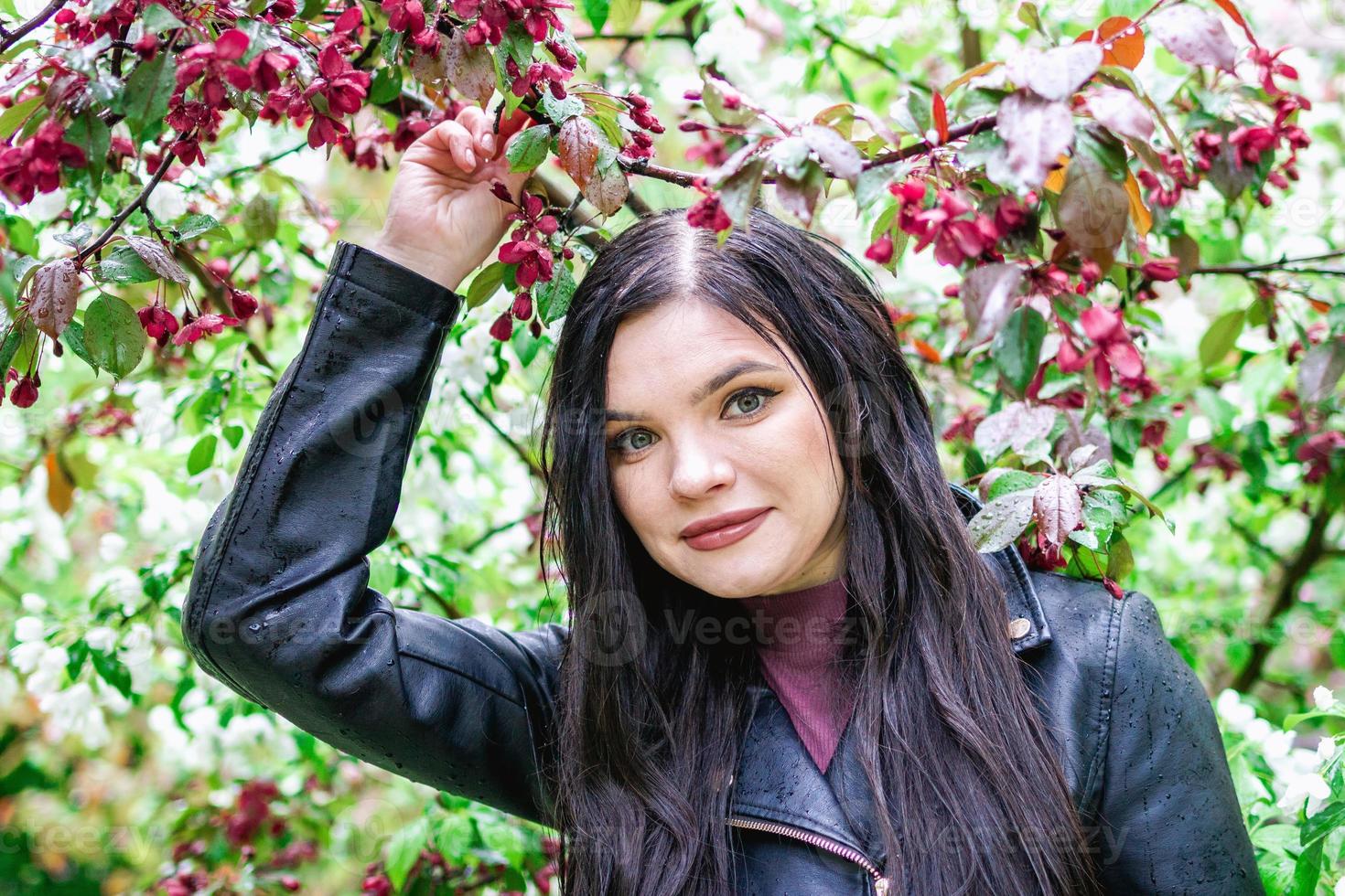 retrato de uma mulher atraente com cabelos castanhos compridos na jaqueta de couro no jardim com flores de macieira flor vermelha e branca e gotas de chuva. foto