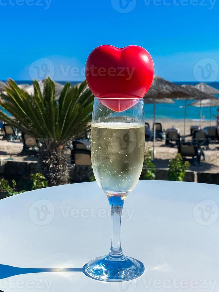 um coração vermelho encontra-se na taça de champanhe com o mar e as palmeiras ao fundo foto